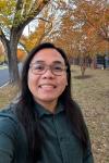 A selfie taken during the Fall season. I'm wearing a dark green long-sleeved shirt, and my background is trees with red and orange leaves on the branches and on the ground.