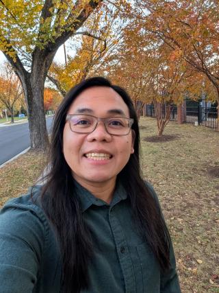 A selfie taken during the Fall season. I'm wearing a dark green long-sleeved shirt, and my background is trees with red and orange leaves on the branches and on the ground.